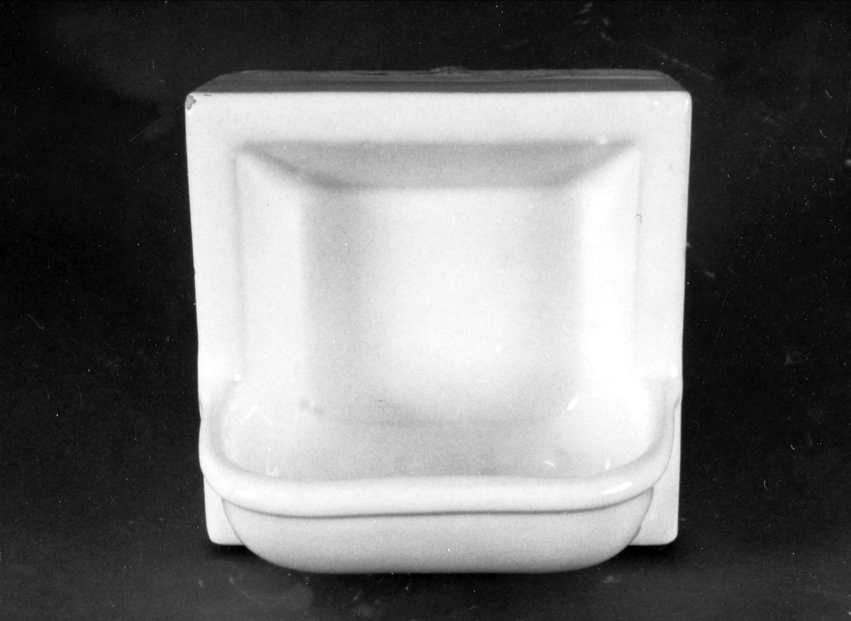 Tjock kakelplatta med tvålkopp, av gulbrännande lera med vit tenn- och blyglasyr. På baksidan 4 fötter, en i varje hörn, stämplad K 1S.