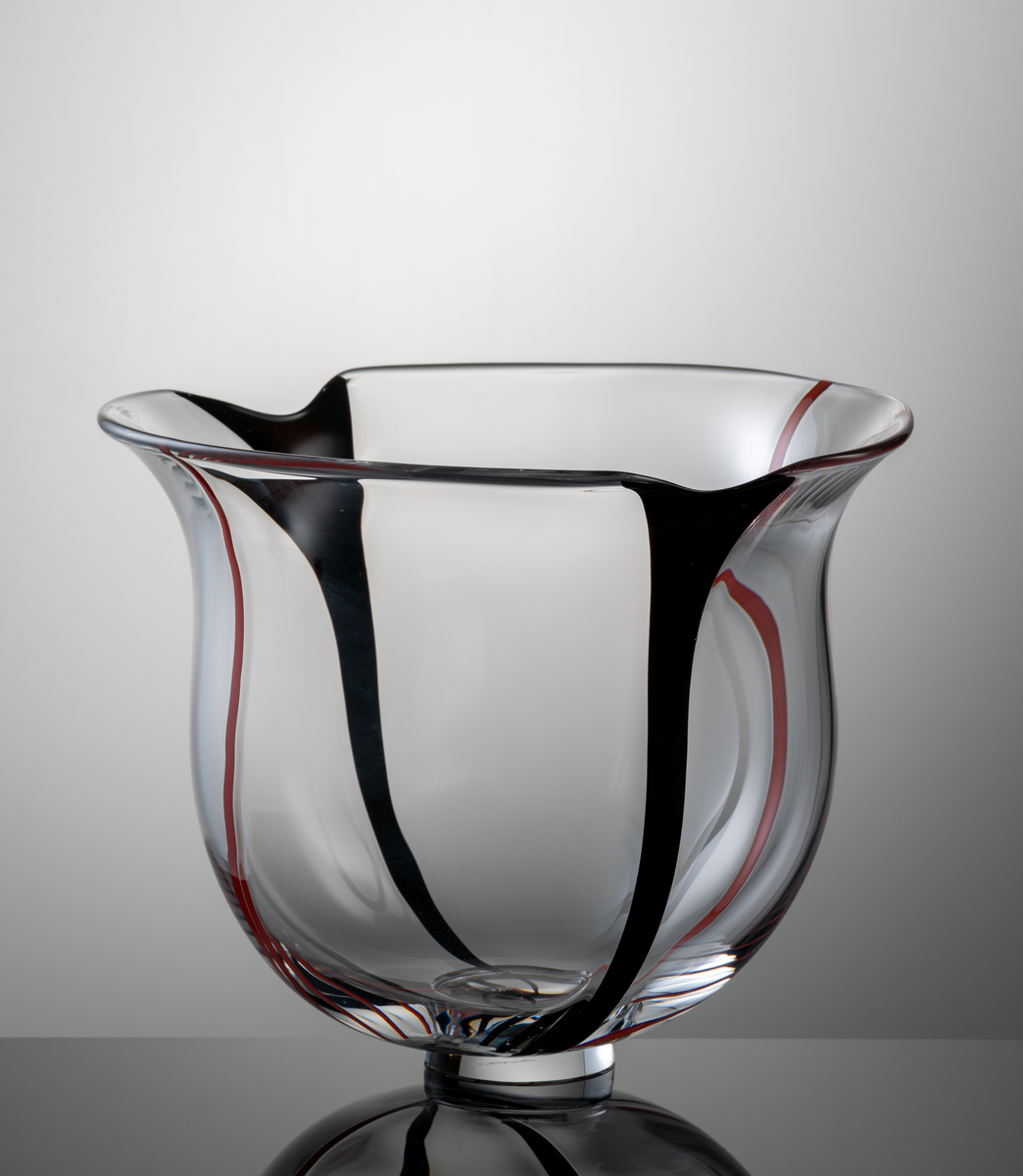 Skål i klart glas, klockformad kupa med två indrag i mynningskanten. I botten en låg fotklack.
Dekor i form av två vertikala svarta resp. röda slingor.
Formgivare: Lars Hellsten.