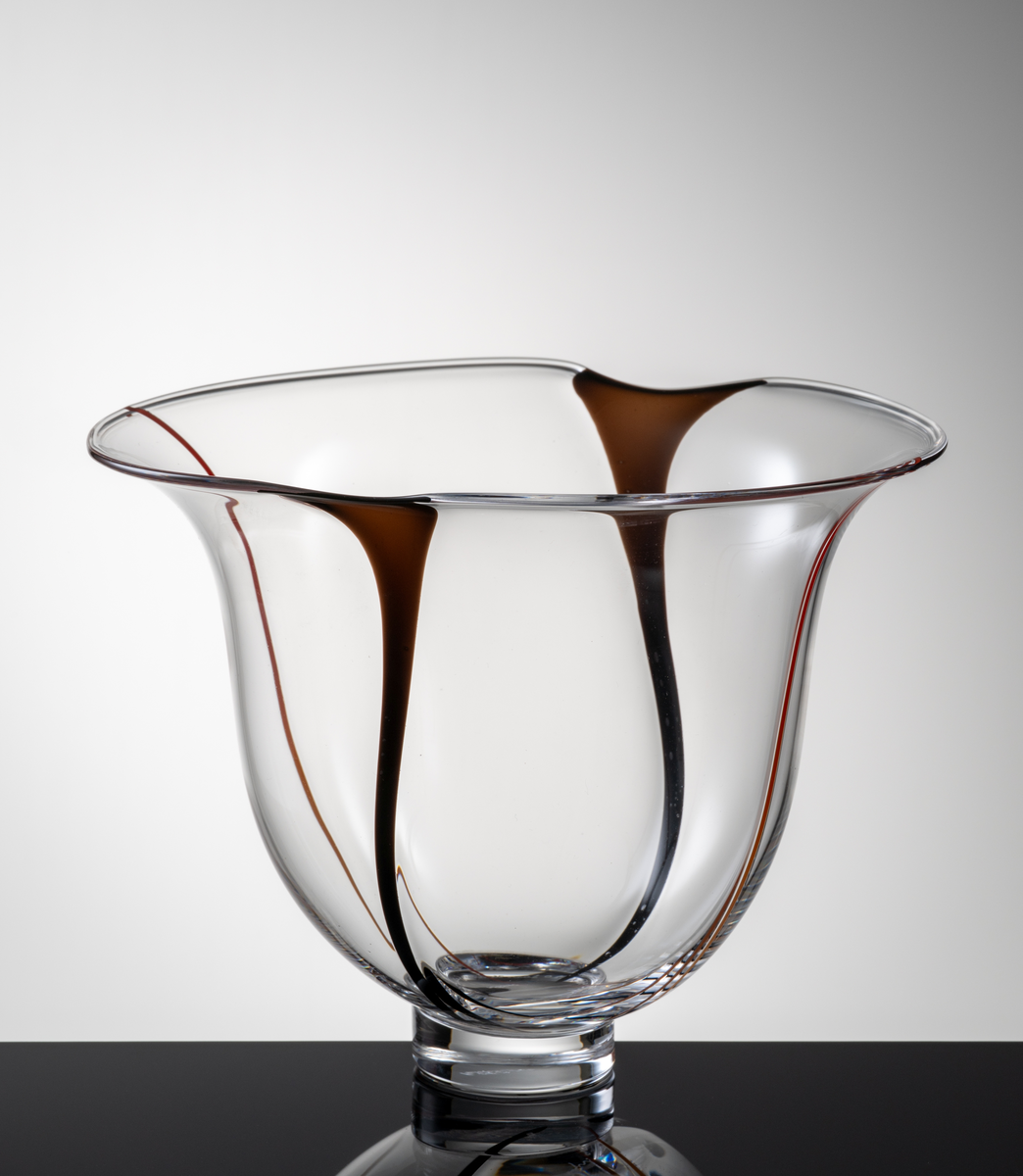 Skål i klart glas, klockformad kupa med två indrag i mynningskanten. I botten en låg fotklack.
Dekor i form av två vertikala svarta resp. röda slingor.
Formgivare: Lars Hellsten.
