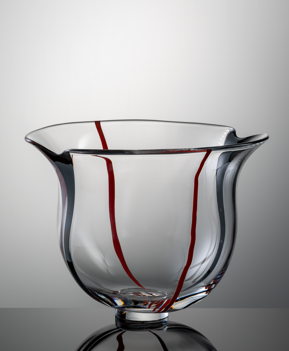 Skål i klart glas, klockformad kupa med två indrag i mynningskanten. I botten en låg fotklack.
Dekor i form av två vertikala svarta resp. röda slingor.
Formgivare: Lars Hellsten.