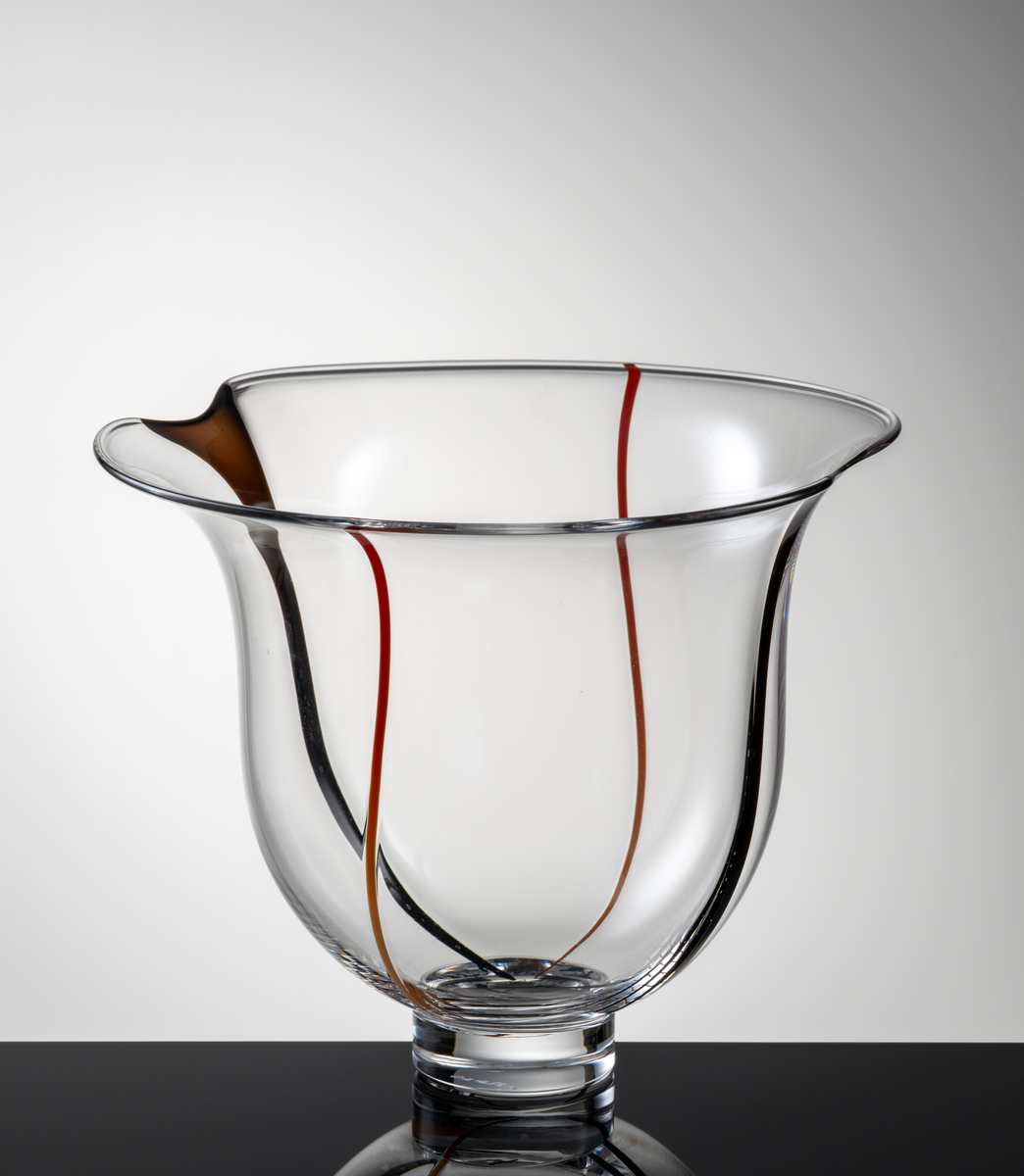 Skål i klart glas, klockformad kupa med två indrag i mynningskanten. I botten en låg fotklack.
Dekor i form av två vertikala svarta resp. röda slingor.
Formgivare: Lars Hellsten.
