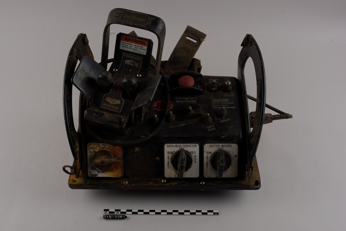 Rektangulær gjenstand. Består av en firkantet base og to halvmåneformede sideplater som er vinklet utover. Sidene er gjennomhullet og fungerer som håndtak. Sideplatene har to metallringer hver, én øverst og én nederst langs halvmåneformen.
Gjenstandens topp og fremside har knapper, brytere og joysticker. Nederst på fronten er det tre vendebrytere, én til ventre og to til høyre. På toppen av gjenstanden syv metall brytere og en rund rød knapp på høyre side. Venstre side har en større spak, utformet som en bøyle, og to mindre spaker med sirkulære hoder. Spakene kan beveges i to retninger, fremover eller bakover.
Gjenstandens høyre side har en ledning. Ledningen er avkappet.
Gjenstandens bakside har en lomme av lær med to stropper av kunststoff.
Gjenstandens front og topp har tekst og klistermerker med tekst. Gjenstandens venstre side har et klistermerke med tekst.