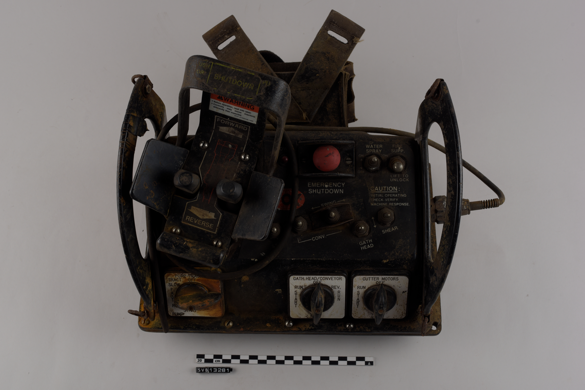 Rektangulær gjenstand. Består av en firkantet base og to halvmåneformede sideplater som er vinklet utover. Sidene er gjennomhullet og fungerer som håndtak. Sideplatene har to metallringer hver, én øverst og én nederst langs halvmåneformen.
Gjenstandens topp og fremside har knapper, brytere og joysticker. Nederst på fronten er det tre vendebrytere, én til ventre og to til høyre. På toppen av gjenstanden syv metall brytere og en rund rød knapp på høyre side. Venstre side har en større spak, utformet som en bøyle, og to mindre spaker med sirkulære hoder. Spakene kan beveges i to retninger, fremover eller bakover.
Gjenstandens høyre side har en ledning. Ledningen er avkappet.
Gjenstandens bakside har en lomme av lær med to stropper av kunststoff.
Gjenstandens front og topp har tekst og klistermerker med tekst. Gjenstandens venstre side har et klistermerke med tekst.