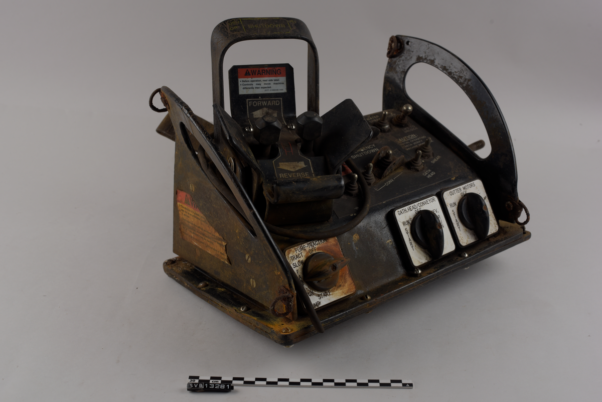 Rektangulær gjenstand. Består av en firkantet base og to halvmåneformede sideplater som er vinklet utover. Sidene er gjennomhullet og fungerer som håndtak. Sideplatene har to metallringer hver, én øverst og én nederst langs halvmåneformen.
Gjenstandens topp og fremside har knapper, brytere og joysticker. Nederst på fronten er det tre vendebrytere, én til ventre og to til høyre. På toppen av gjenstanden syv metall brytere og en rund rød knapp på høyre side. Venstre side har en større spak, utformet som en bøyle, og to mindre spaker med sirkulære hoder. Spakene kan beveges i to retninger, fremover eller bakover.
Gjenstandens høyre side har en ledning. Ledningen er avkappet.
Gjenstandens bakside har en lomme av lær med to stropper av kunststoff.
Gjenstandens front og topp har tekst og klistermerker med tekst. Gjenstandens venstre side har et klistermerke med tekst.