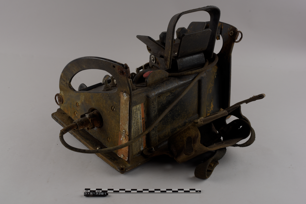 Rektangulær gjenstand. Består av en firkantet base og to halvmåneformede sideplater som er vinklet utover. Sidene er gjennomhullet og fungerer som håndtak. Sideplatene har to metallringer hver, én øverst og én nederst langs halvmåneformen.
Gjenstandens topp og fremside har knapper, brytere og joysticker. Nederst på fronten er det tre vendebrytere, én til ventre og to til høyre. På toppen av gjenstanden syv metall brytere og en rund rød knapp på høyre side. Venstre side har en større spak, utformet som en bøyle, og to mindre spaker med sirkulære hoder. Spakene kan beveges i to retninger, fremover eller bakover.
Gjenstandens høyre side har en ledning. Ledningen er avkappet.
Gjenstandens bakside har en lomme av lær med to stropper av kunststoff.
Gjenstandens front og topp har tekst og klistermerker med tekst. Gjenstandens venstre side har et klistermerke med tekst.