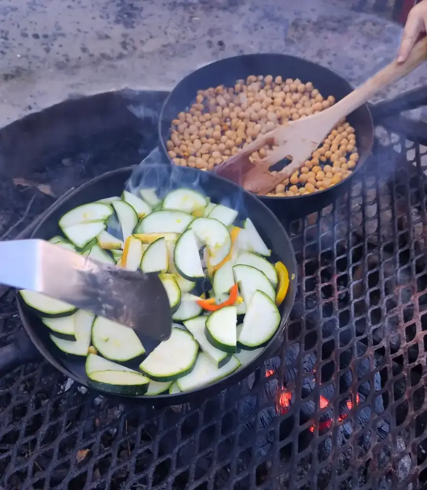 Två stekpannor över en öppen eld, som steker olika grönsaker.