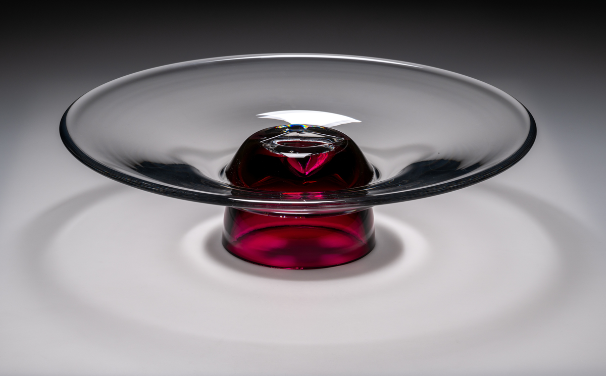 Formgivare: Lars Hellsten. Fat/skål på fot. Klockformad övre del med utvikt brätte. Rosa fot i form av en omvänd rund skål. Fatet är nedtryckt över foten. Sammanfogade varmt i hyttan.