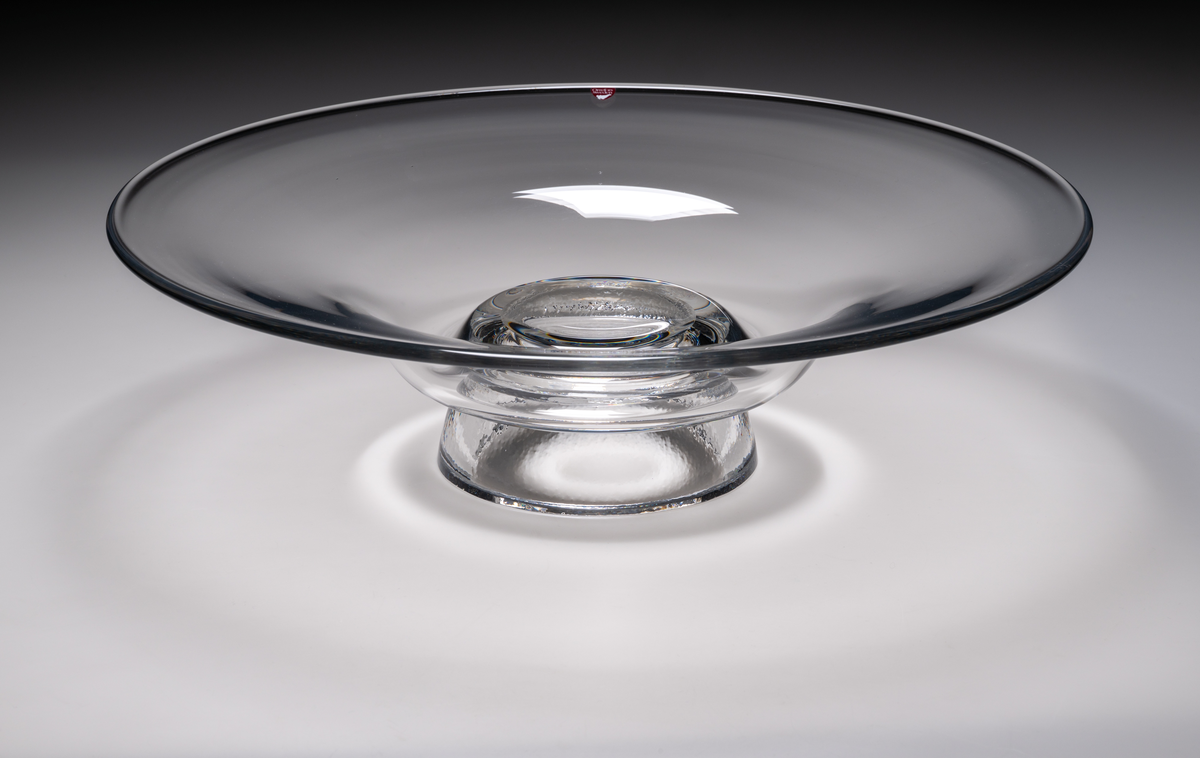 Formgivare: Lars Hellsten. Fat/skål på fot i ofärgat glas. Klockformad övre del med utvikt brätte. Fot i form av en omvänd rund skål. Fatet är nedtryckt över foten. Sammanfogade varmt i hyttan.