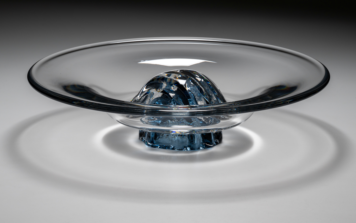 "Havsblomma" Formgivare: Lars Hellsten. Skål på fot. Klockformad övre del i ofärgat glas. Fot i form av en blå ”blomma” med breda räfflor. Fatet är nedtryckt över foten. Sammanfogade varmt i hyttan.