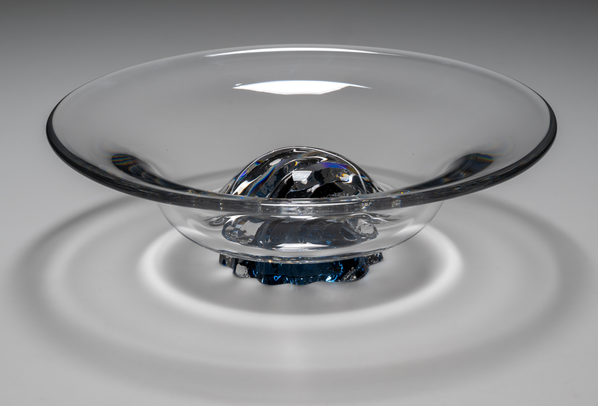 "Havsblomma" Formgivare: Lars Hellsten. Skål på fot. Klockformad övre del i ofärgat glas. Fot i form av en blå ”blomma” med breda räfflor. Fatet är nedtryckt över foten. Sammanfogade varmt i hyttan.