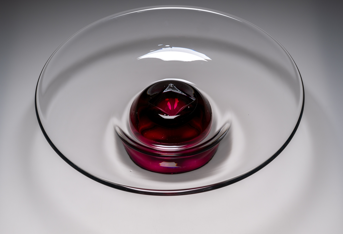 Formgivare: Lars Hellsten. Fat/skål på fot. Klockformad övre del med utvikt brätte. Rosa fot i form av en omvänd rund skål. Fatet är nedtryckt över foten. Sammanfogade varmt i hyttan.