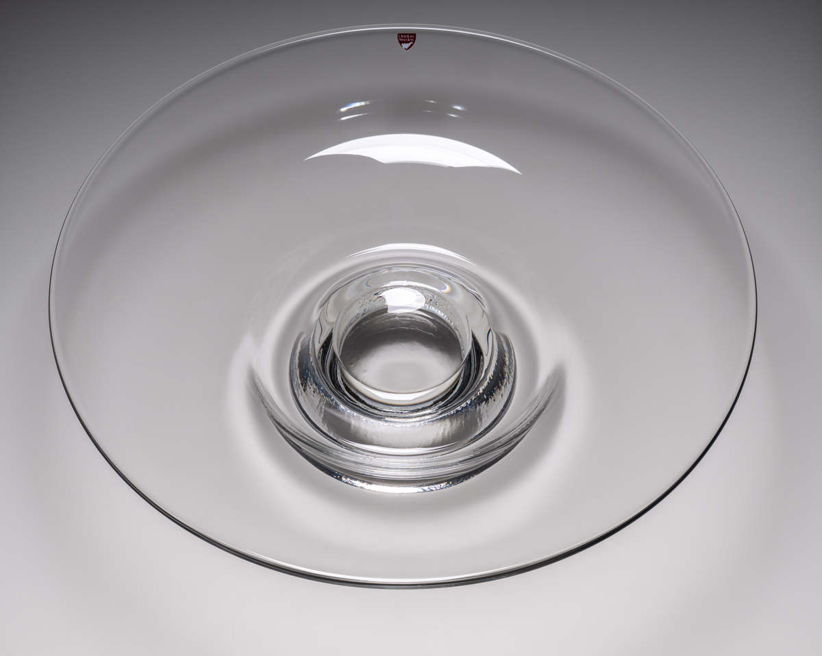 Formgivare: Lars Hellsten. Fat/skål på fot i ofärgat glas. Klockformad övre del med utvikt brätte. Fot i form av en omvänd rund skål. Fatet är nedtryckt över foten. Sammanfogade varmt i hyttan.