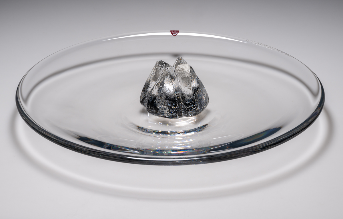 Formgivare: Lars Hellsten. Fat i klarglas, med rund form. I fatets mitt, en gjuten, skrovlig ”blomma” i klarglas.