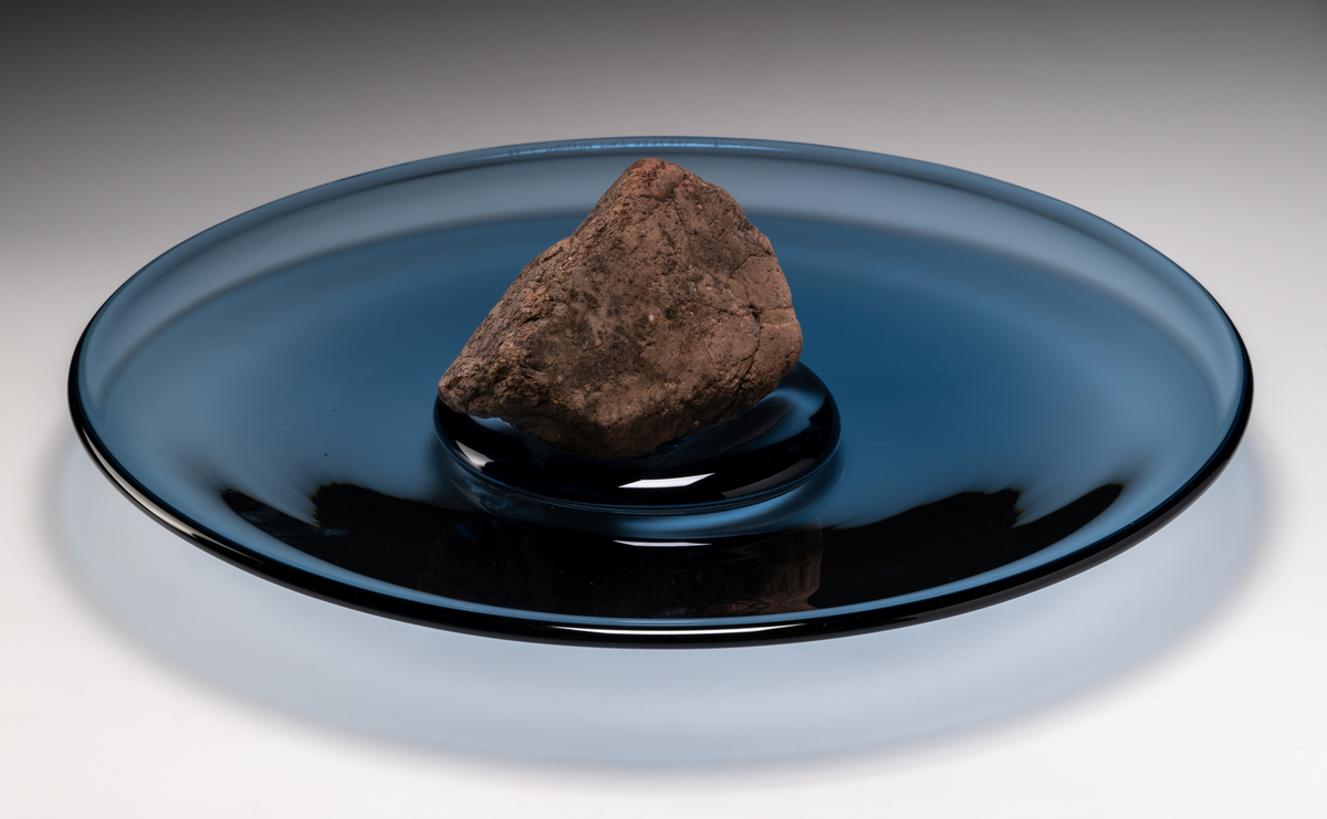 Formgivare: Lars Hellsten. Fat i midnattsblått glas, med rund form. I fatets mitt, ett pålagt fält av klarglas, vari en sten är placerad.