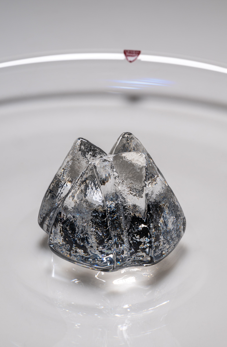 Formgivare: Lars Hellsten. Fat i klarglas, med rund form. I fatets mitt, en gjuten, skrovlig ”blomma” i klarglas.