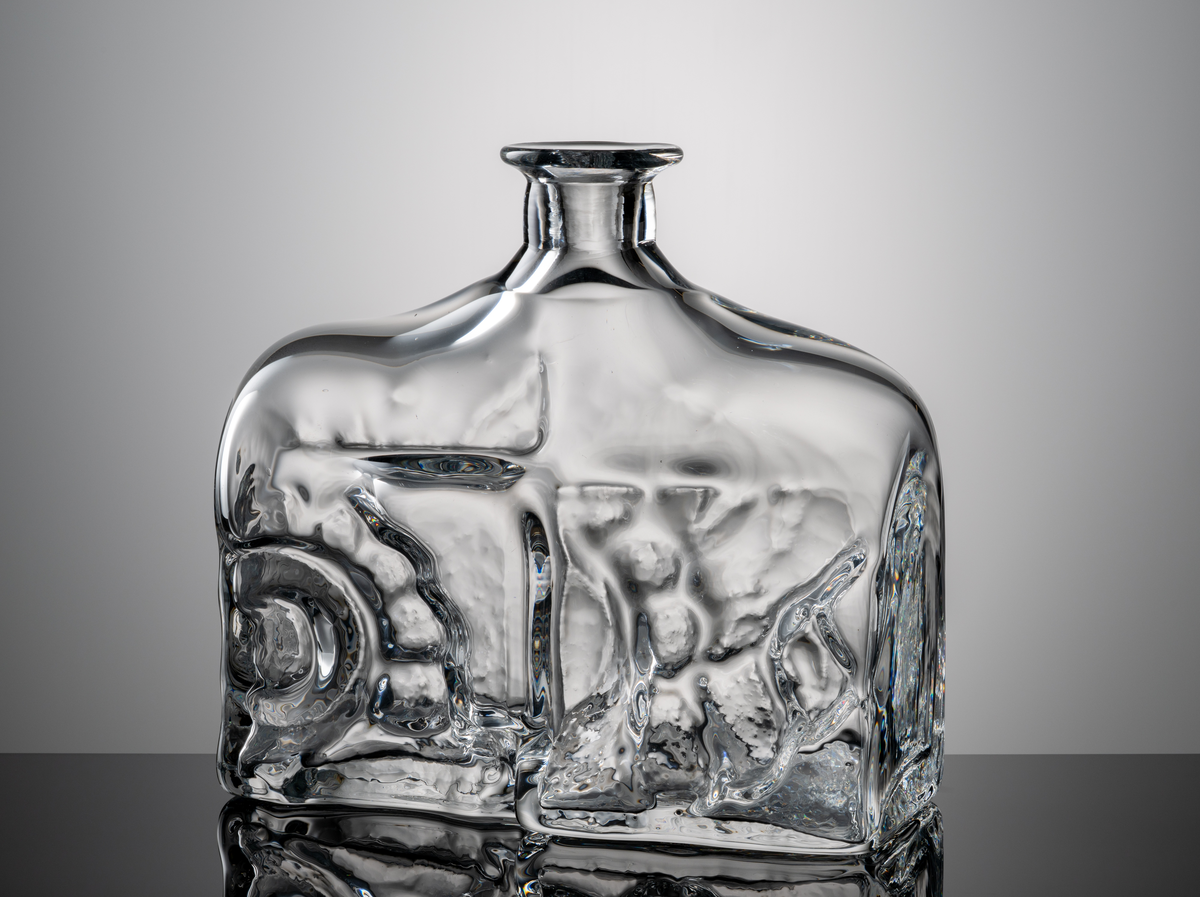 Formgivare: Lars Hellsten. Flaska i klarglas med rektangulär kropp och kort hals. Oregelbundet reliefmönster på främre väggen.