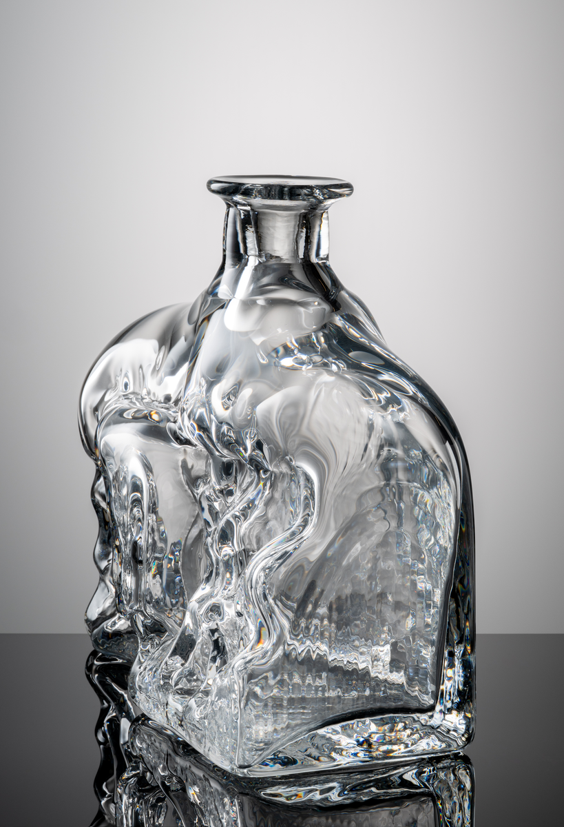 Formgivare: Lars Hellsten. Flaska i klarglas med rektangulär kropp och kort hals. Oregelbundet reliefmönster på främre väggen.