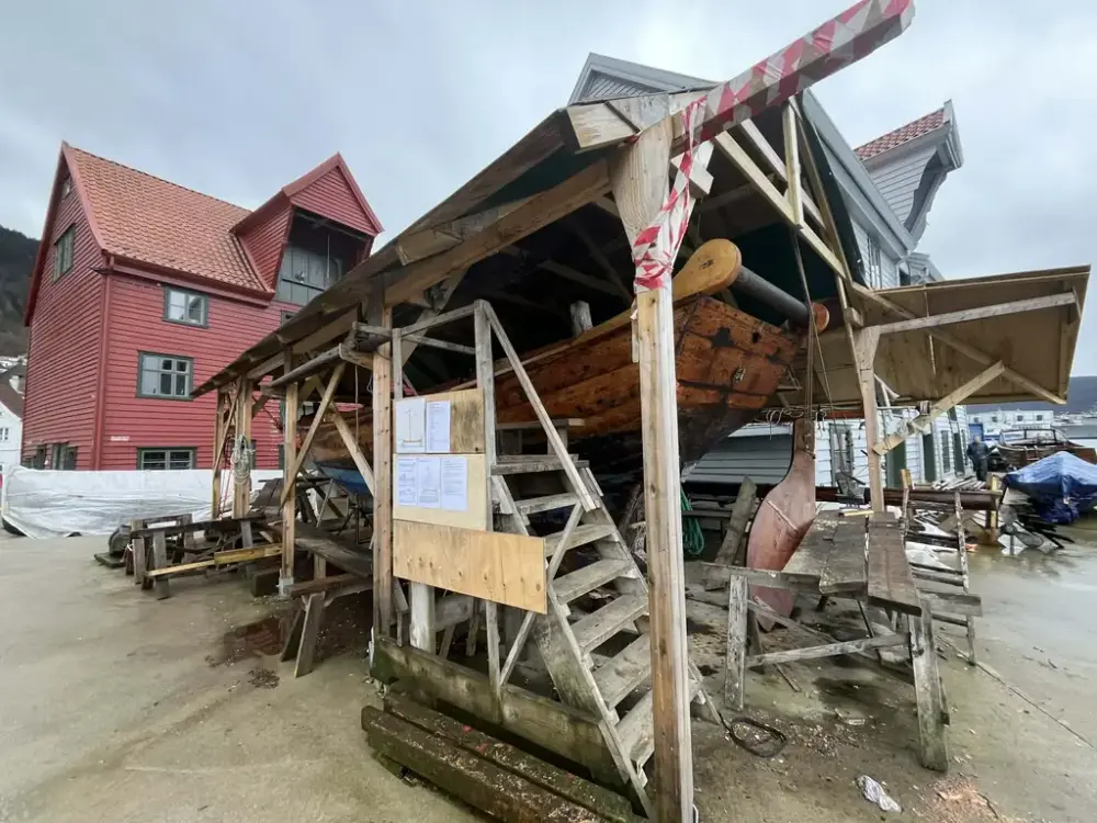 Vi ser en gavlbåt under restaurering. Den står under et tak. 
