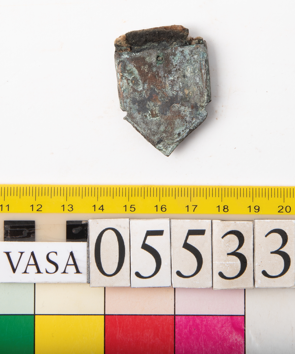 Remändebeslag av koppar med kvarsittande fragmentariska rester av läder. Lädret har fästs med stift genom beslaget. Beslaget är ärgat men relativt välbevarat.