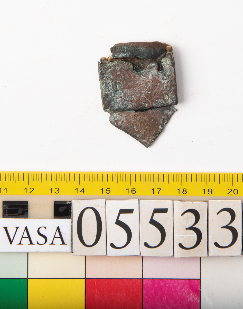 Remändebeslag av koppar med kvarsittande fragmentariska rester av läder. Lädret har fästs med stift genom beslaget. Beslaget är ärgat men relativt välbevarat.