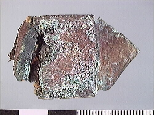 Remändebeslag av koppar med kvarsittande fragmentariska rester av läder. Lädret har fästs med stift genom beslaget. Beslaget är ärgat men relativt välbevarat.