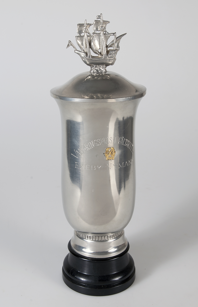 Bägare av tenn med lock, på svartbetsad träfot, med graverad text: Vandringspris i fältskytte Ekeby - Nymans, och med mindre stil: 1954 Nymans, 1955 Nymans. Locket försett med ett segelfartyg.