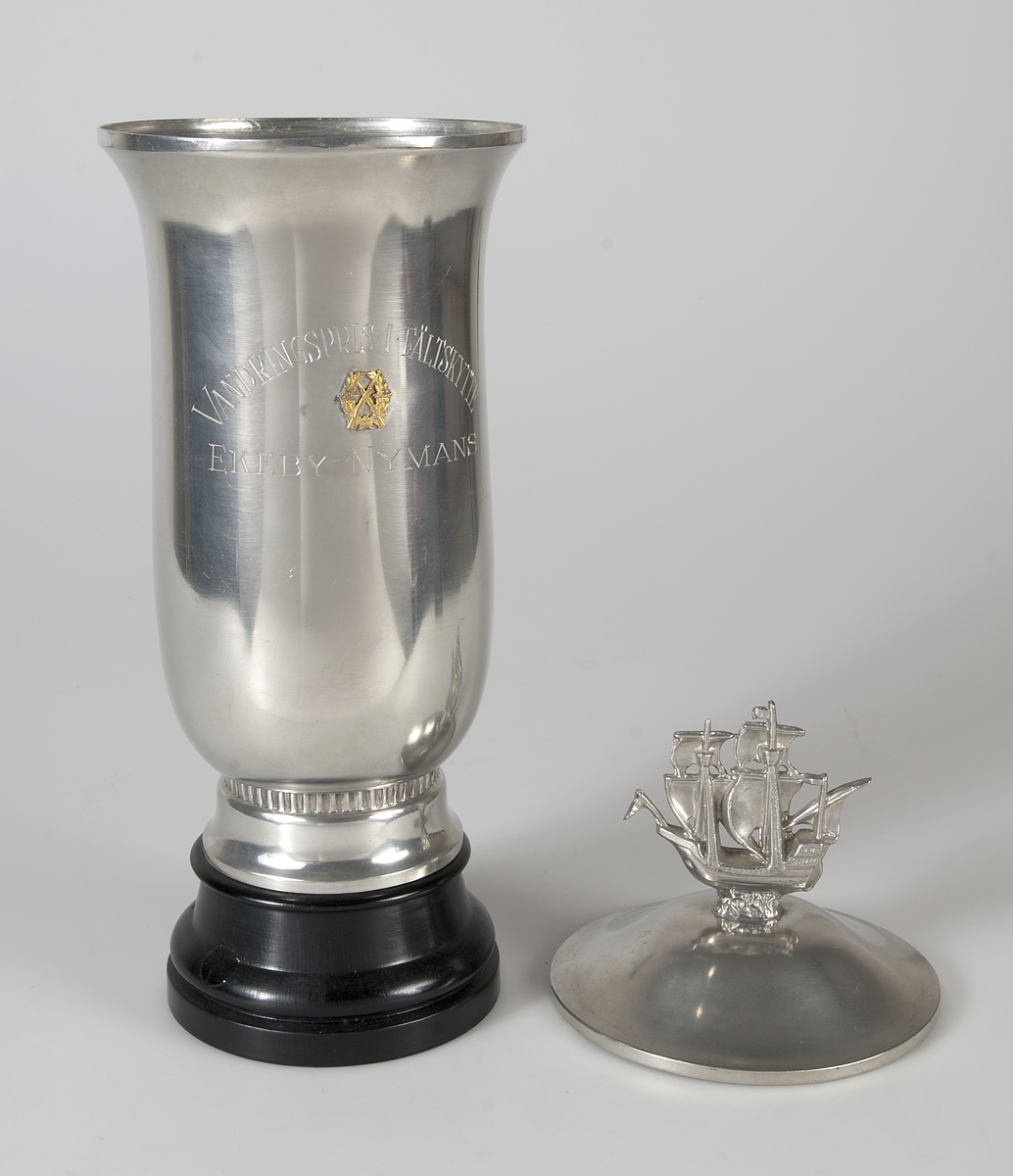 Bägare av tenn med lock, på svartbetsad träfot, med graverad text: Vandringspris i fältskytte Ekeby - Nymans, och med mindre stil: 1954 Nymans, 1955 Nymans. Locket försett med ett segelfartyg.