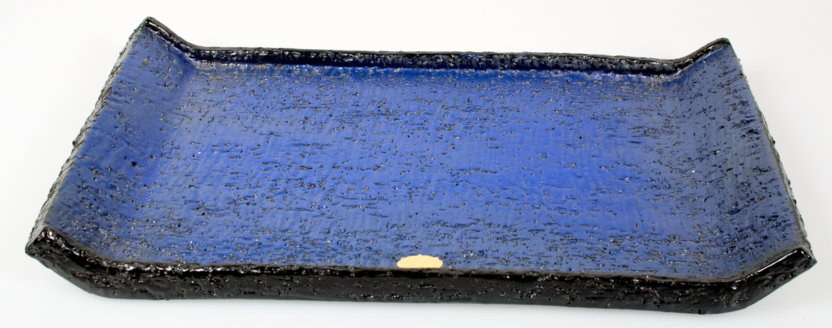 Platt fyrkantigt fat av grov chamottlera med sluttande kant på kortsidorna och uppvikt rak kant på långsidorna. Blåglaserat. Upsala-Ekebys gula pappersmärke. Saknar tillverkarstämplar.