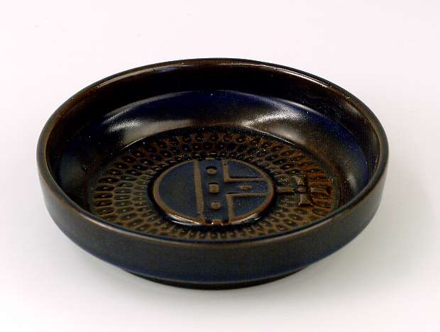 Askfat av glaserat brunt/mörkblått lergods. Upplandsvapnet på botten. På undersidan stämplat: UE SWEDEN.