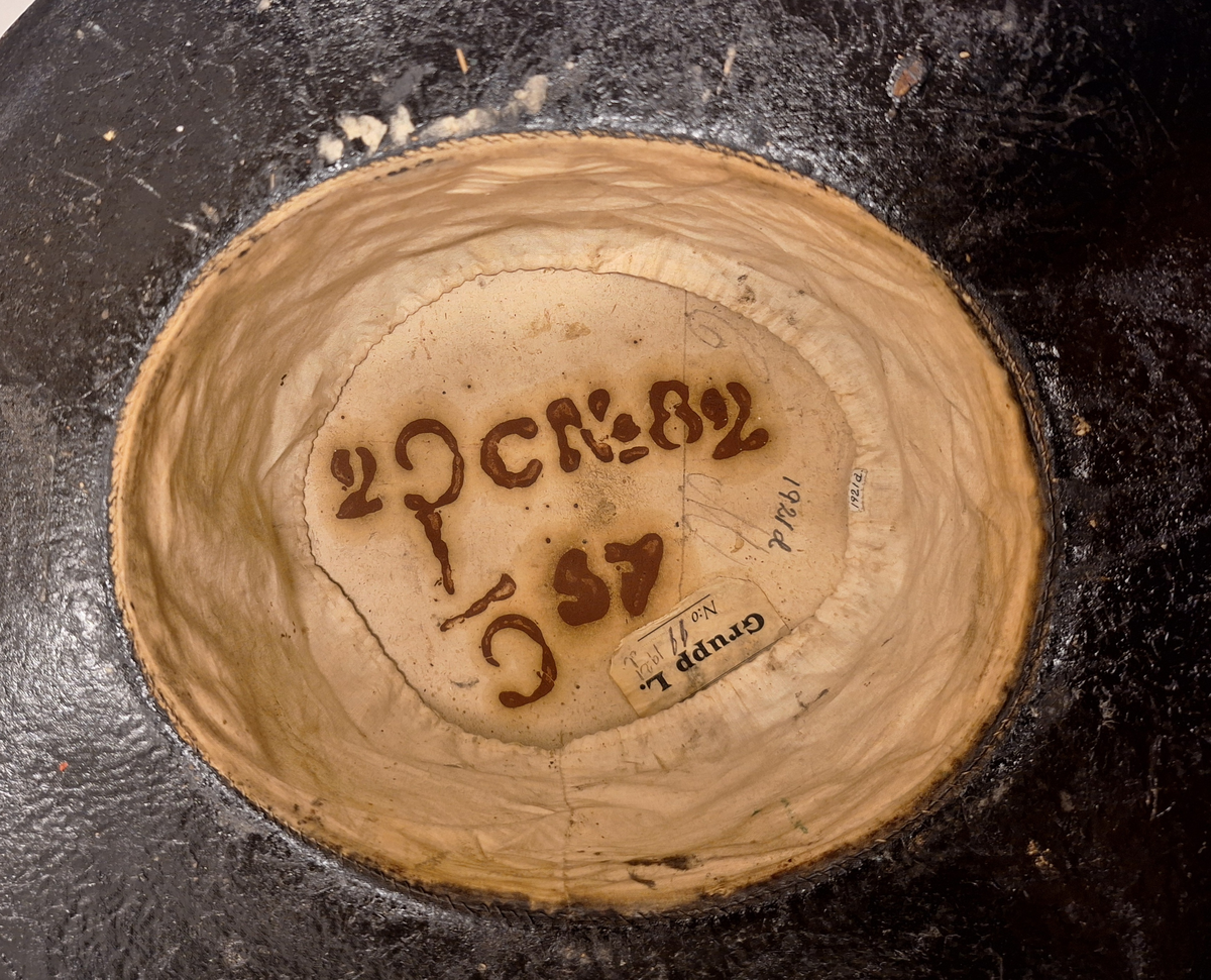 Svartlackerad papphatt, halvhög cylinder med breda brätten. Sidenband med texten ”K.Skärgårdsartilleriet” i guldtryck. Fodrad med vitt linne.