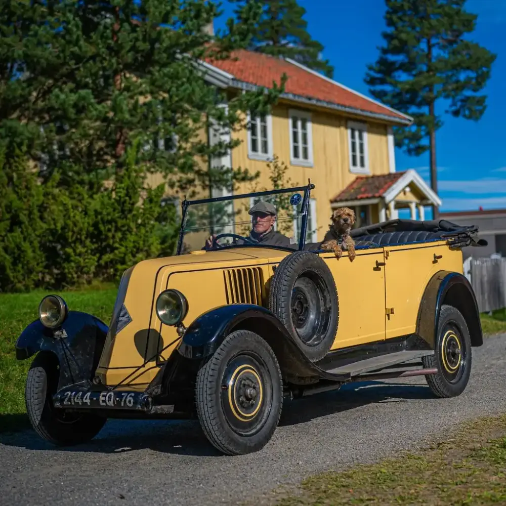 Gul og svart veteranbil med sjåfør og liten hund foran et historisk trehus på museum.