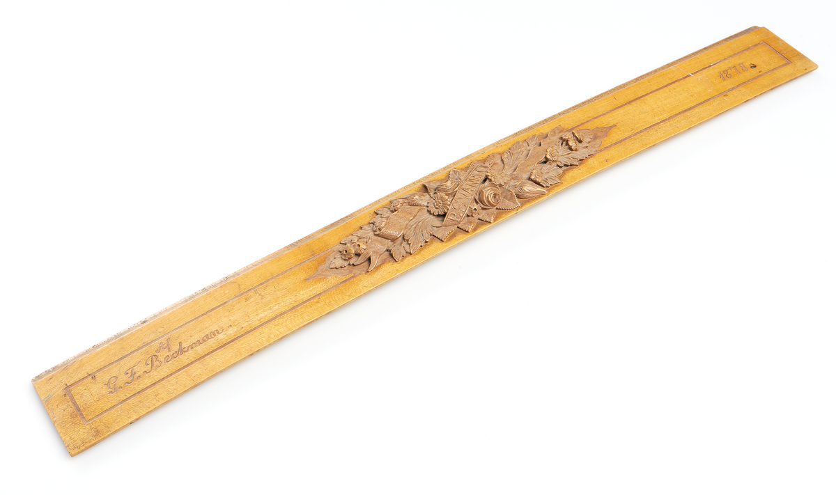 Linjal av ljust trä. Märkt "Af G F Beckman" "1878". Med snidade blommor, blad och banderoll på mittten, märkt P Svanbeck. 

Insrivet i huvudbok år 1969.