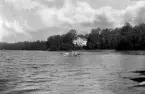 Herrgård i lövskog intill vattendrag, samt fyra kvinnor i en eka.
