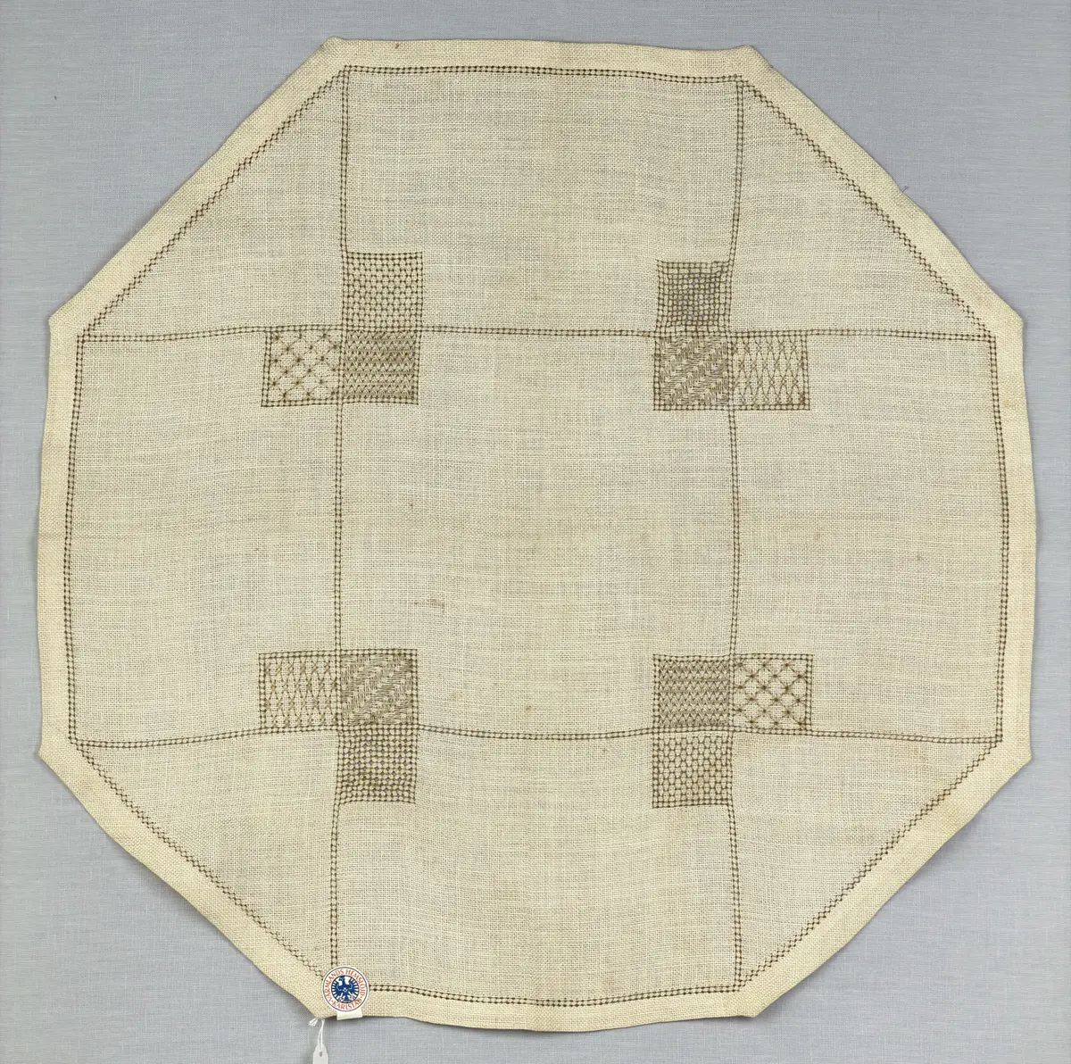 Åttkantig duk gjord av linneväv med 10tr/cm i tuskaft och broderi utfört med lingarn i sex olika bottensömmar. Dukens kanter har en 16mm bred dubbelvikt fåll runt om fastsydd med kaststygn samt en myrgångs- söm.