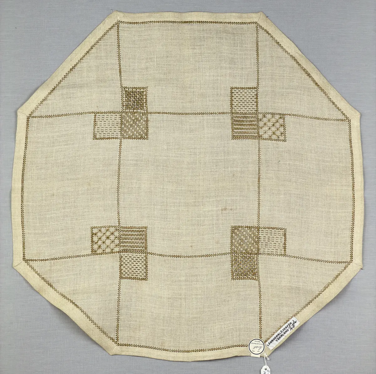 Åttkantig duk gjord av linneväv med 10tr/cm i tuskaft och broderi utfört med lingarn i sex olika bottensömmar. Dukens kanter har en 16mm bred dubbelvikt fåll runt om fastsydd med kaststygn samt en myrgångs- söm.