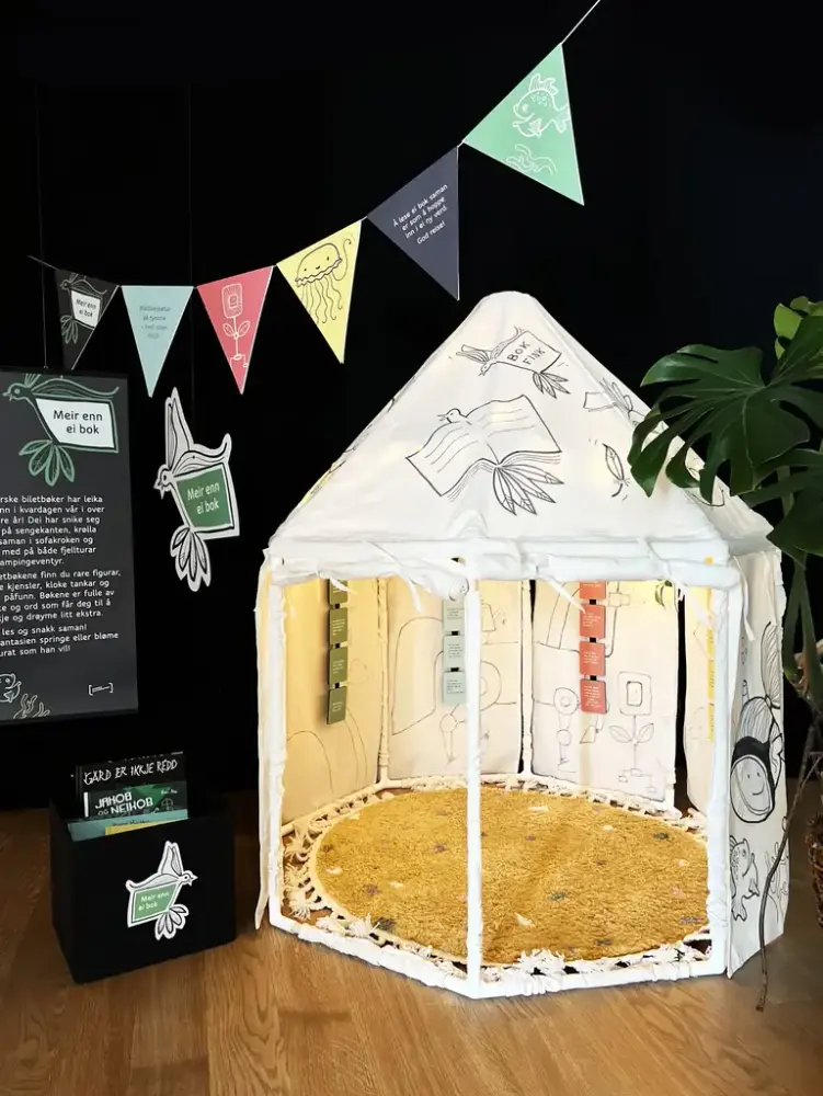Eit kvitt telt med illustrasjonar står under nokre fargerike vimplar. Ved sidan av står det ein svat boks med bøker i og over den eit skilt med tekst.