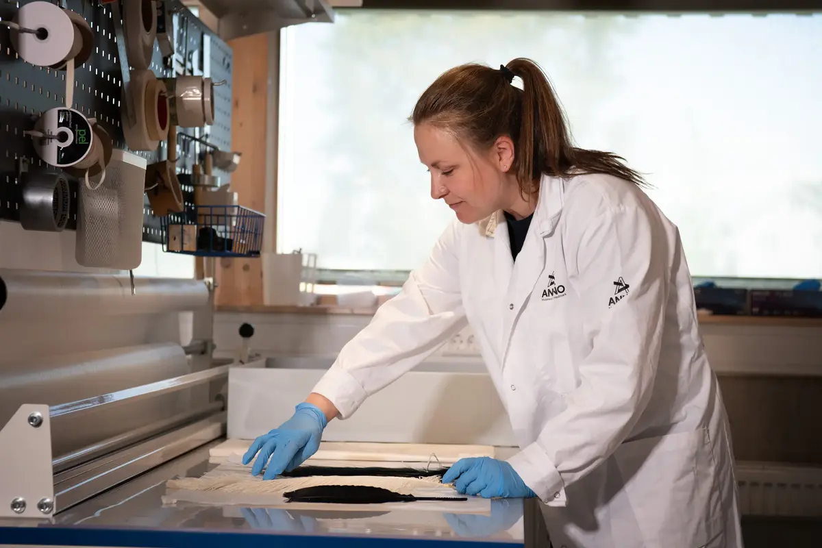 Bilde av magasinforvalter ved Anno bevaringssenter, Toril Skaaraas Hofseth. Hun står ved et bord og ser på og berører en liten, avlang, mørk gjenstand. 