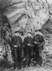 Tre menn med sigarettar og luer framfor ein fjellvegg.
