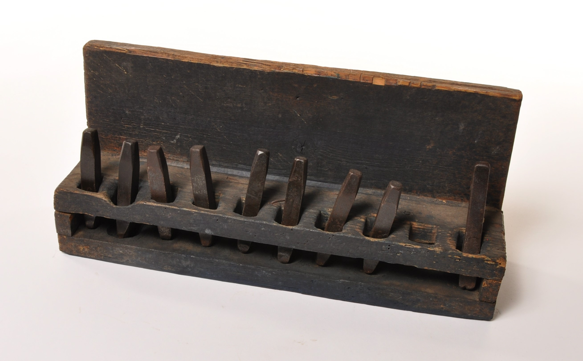 Åpen hylle for plassering av håndpunsler. Kasse av tre, med ryggstykke og dobbelt hylle med en rad kvadratiske hull til 10 stk løse merkejern. Jernene er merket b-k, hvorav j mangler.