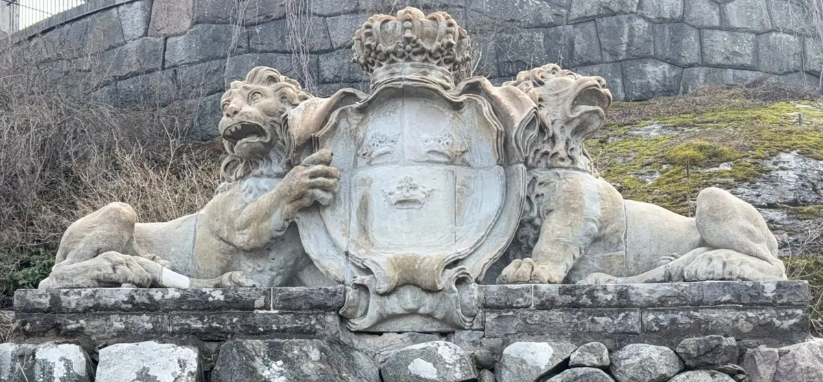 Fasadskulptur av sandsten. Två liggande lejon på ömse sidor om en krönt sköld med tre kronor (lilla riksvapnet). Sockelstenar i kalksten. 