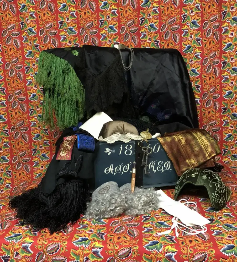 Öppnad kista med kläder i olika färger och en mönstrad röd tapet i bakgrunden.