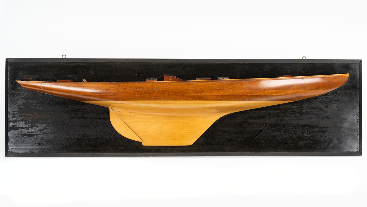 Halvmodell av segelyachten EBE 150 kvm, byggd 1920. Modellen byggd i block av mahogny, mörk sida, ljus botten, rorkult, sittbrunn, tre skylight och två kappluckor, monterad på svart platta.