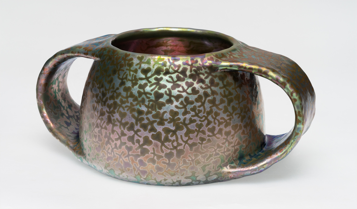 Vase i fajanse, hvor overflaten er dekorert med luster. Halvkuleformet korpus, flankert av to kraftige halvsirkelformede hanker. Overflaten domineres av skimrende horisontale fargepartier i rød, grønn og blå med abstraherte kløverfremstillinger.