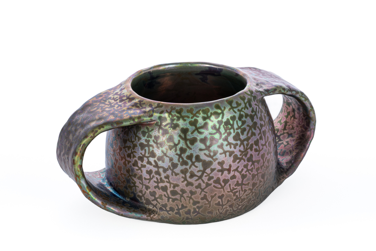 Vase i fajanse, hvor overflaten er dekorert med luster. Halvkuleformet korpus, flankert av to kraftige halvsirkelformede hanker. Overflaten domineres av skimrende horisontale fargepartier i rød, grønn og blå med abstraherte kløverfremstillinger.