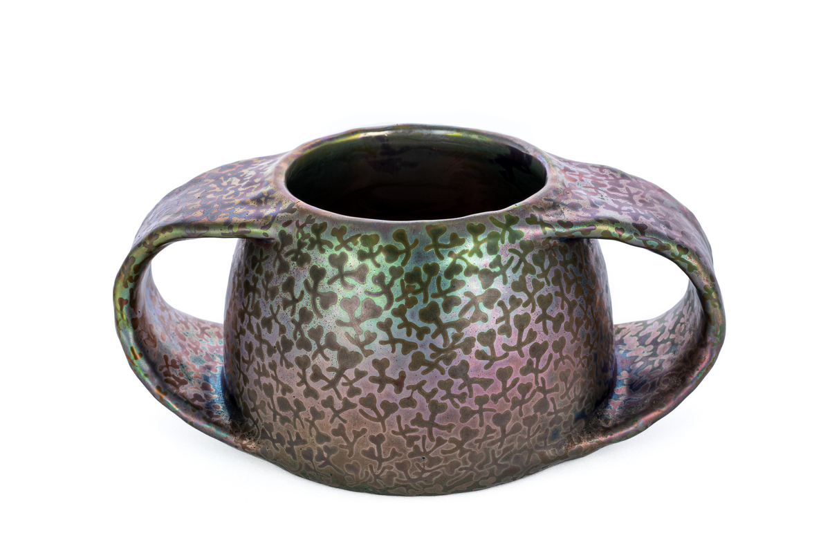 Vase i fajanse, hvor overflaten er dekorert med luster. Halvkuleformet korpus, flankert av to kraftige halvsirkelformede hanker. Overflaten domineres av skimrende horisontale fargepartier i rød, grønn og blå med abstraherte kløverfremstillinger.