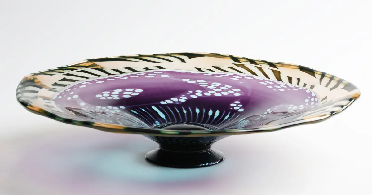 "Monogram" Formgivare: Eva Englund. Graalfat med svart låg fot och oregelbundet brätte. Mönsterdekor i violett och ljusblå nyans i fatets mitt och blekrosa och svart bård runt brättet.