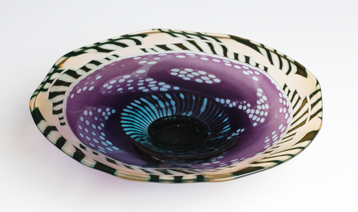 "Monogram" Formgivare: Eva Englund. Graalfat med svart låg fot och oregelbundet brätte. Mönsterdekor i violett och ljusblå nyans i fatets mitt och blekrosa och svart bård runt brättet.