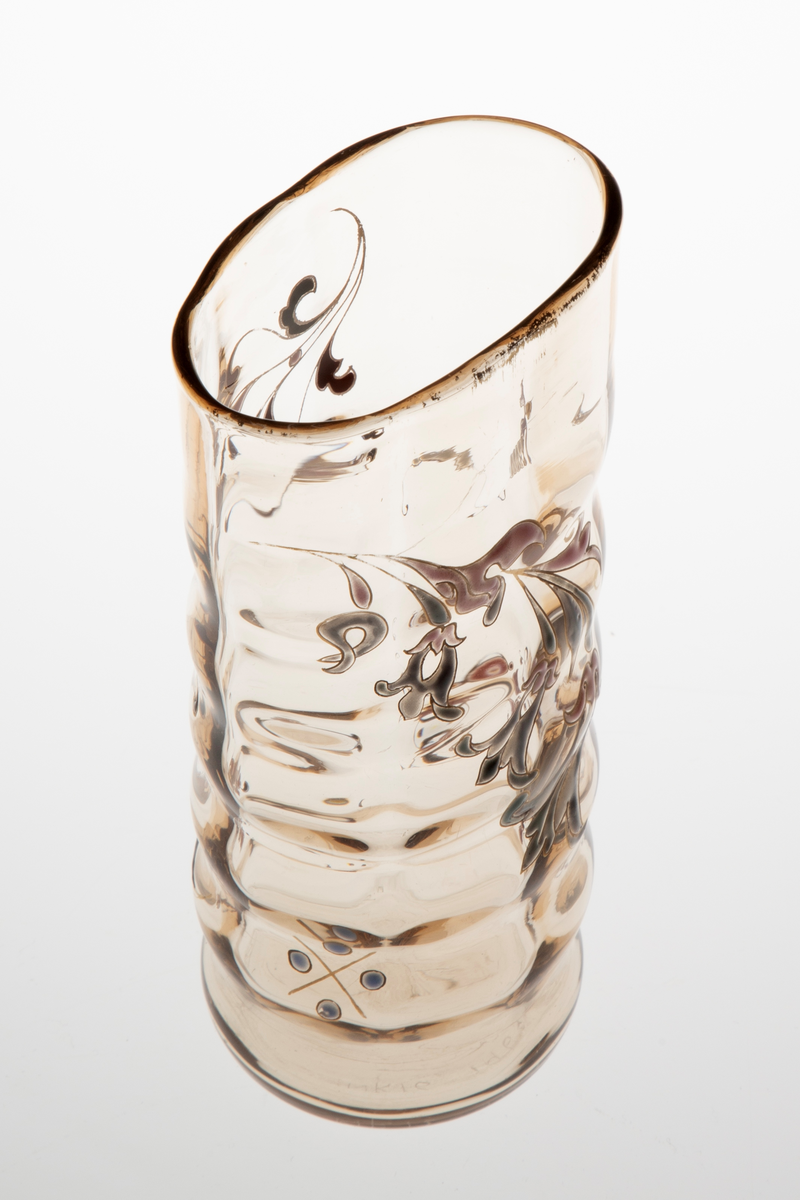 Begerformet vase i gjennomskinnelig glass med gulbrun fargetone. Lett bølgende vegger, som skaper en dynamisk bevegelse i vasens utforming. Sirkulær fot, som gradvis glir over i en ovalform på øvre del av korpus. Vasen avsluttes med en forgyllet munningskant. Korpus er dekorert med organiske motiver i emaljemaling. På den ene siden er blomster og blomstergrener primært fremstilt i burgund og sort, mens på den andre siden er de holdt i grått og rosa. Emaljemalingen innrammes av forgylte konturlinjer.