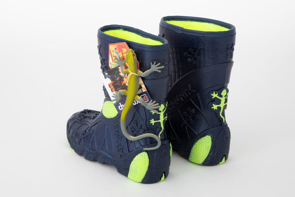 Barnestøvler i metallisk blå PVC med syntetisk pelsfôr. Gulgrønne detaljer og fôr. Støvlene er mønsteret med blant annet små gekkoer og merkenavnet. Liten gekko-lekefigur og originaleske følger med.