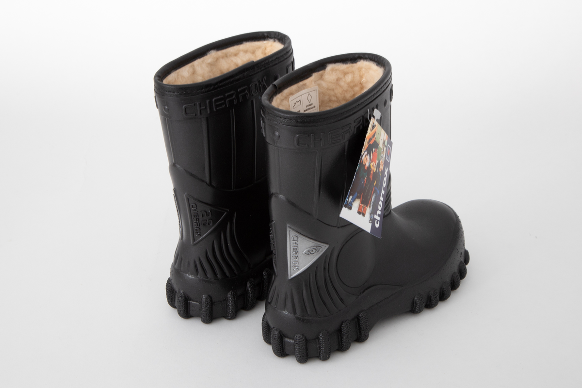 Barnestøvler i svart PVC med kremfarget syntetisk pelsfôr. Enkel stripedekor på skaftet. Merkenavn øverst rundt skaftet og på trekantede detaljer på baksiden. Originaleske følger med.